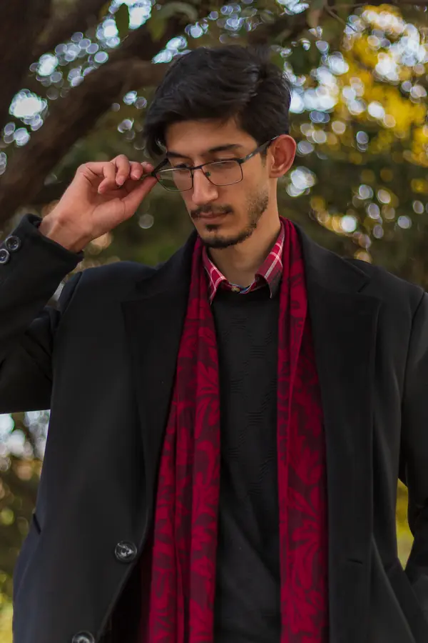 Hadi Jafari in a black coat and red scarf, adjusting his glasses under autumn trees